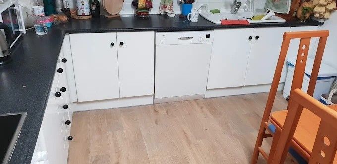 Una cocina con armarios blancos, lavavajillas y suelo de madera. Hay dos sillas naranjas.