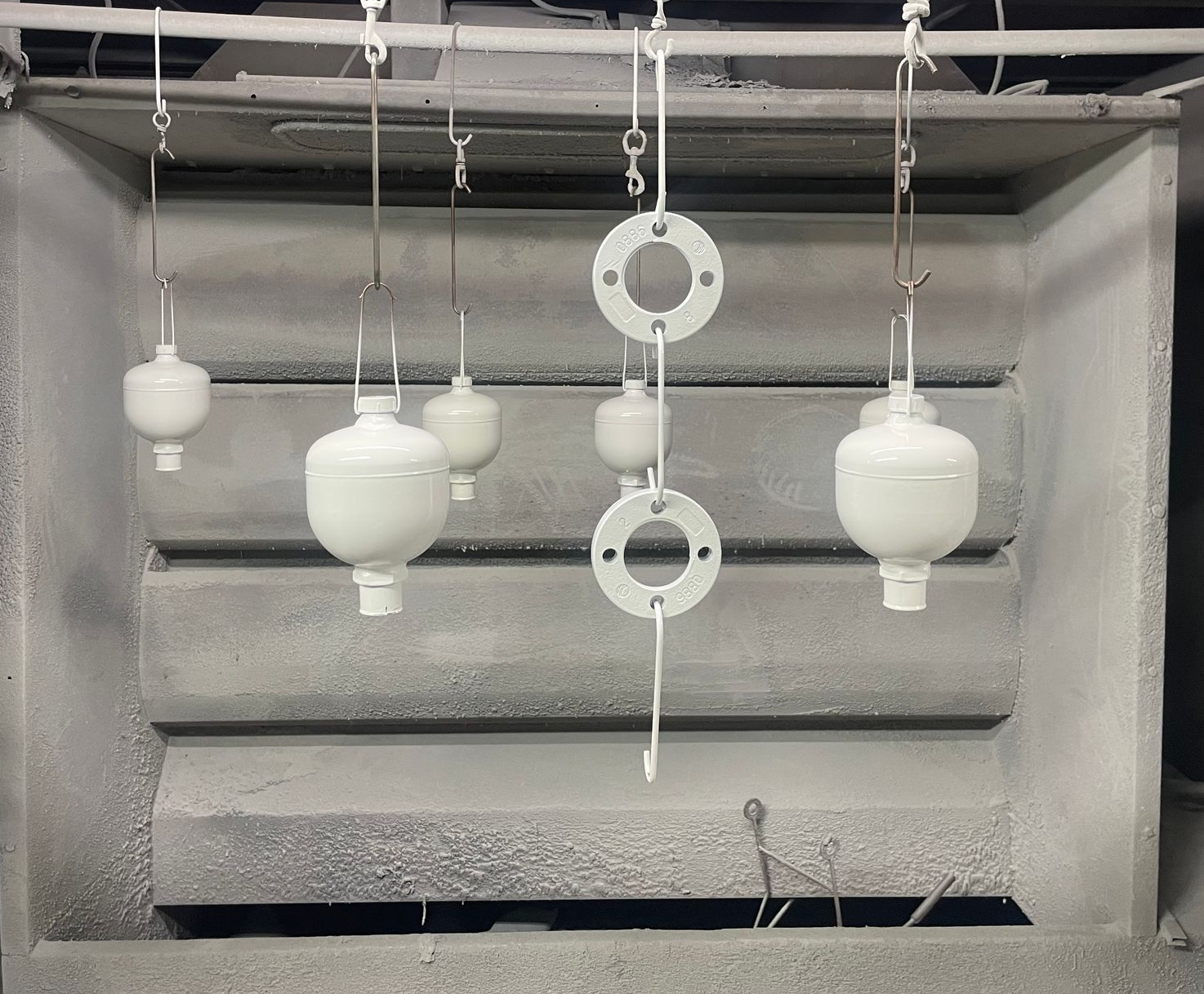 Des pièces métalliques peintes en blanc sont suspendues à des crochets à l'intérieur d'une cabine de peinture.