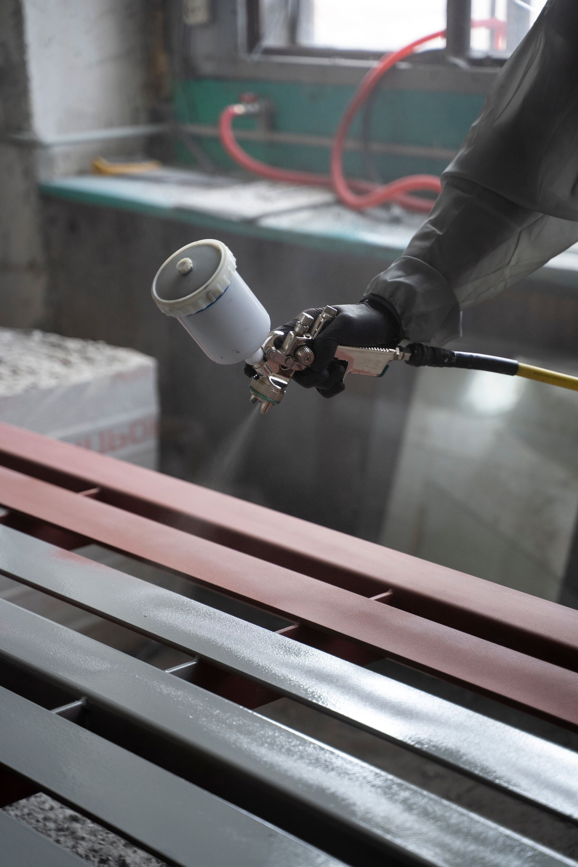 Une personne pulvérise de la peinture rouge sur des poutres métalliques à l'aide d'un pistolet à peinture, à l'intérieur.