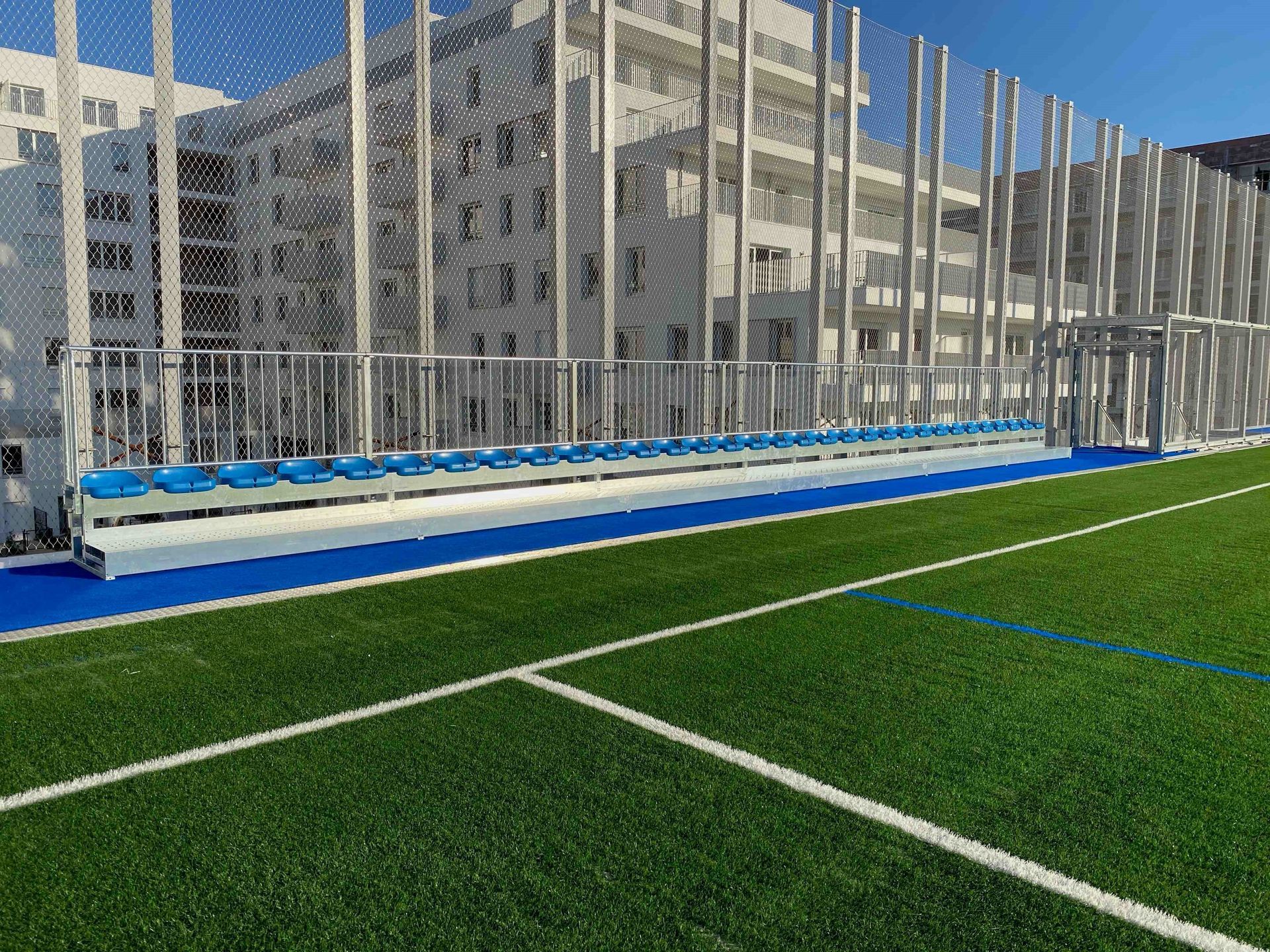 Un campo de fútbol con una valla y asientos azules delante de un edificio.