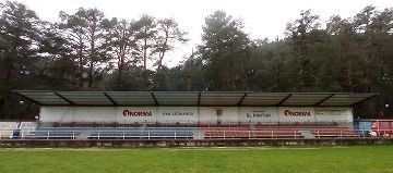 Un campo de fútbol con una grada y árboles al fondo.