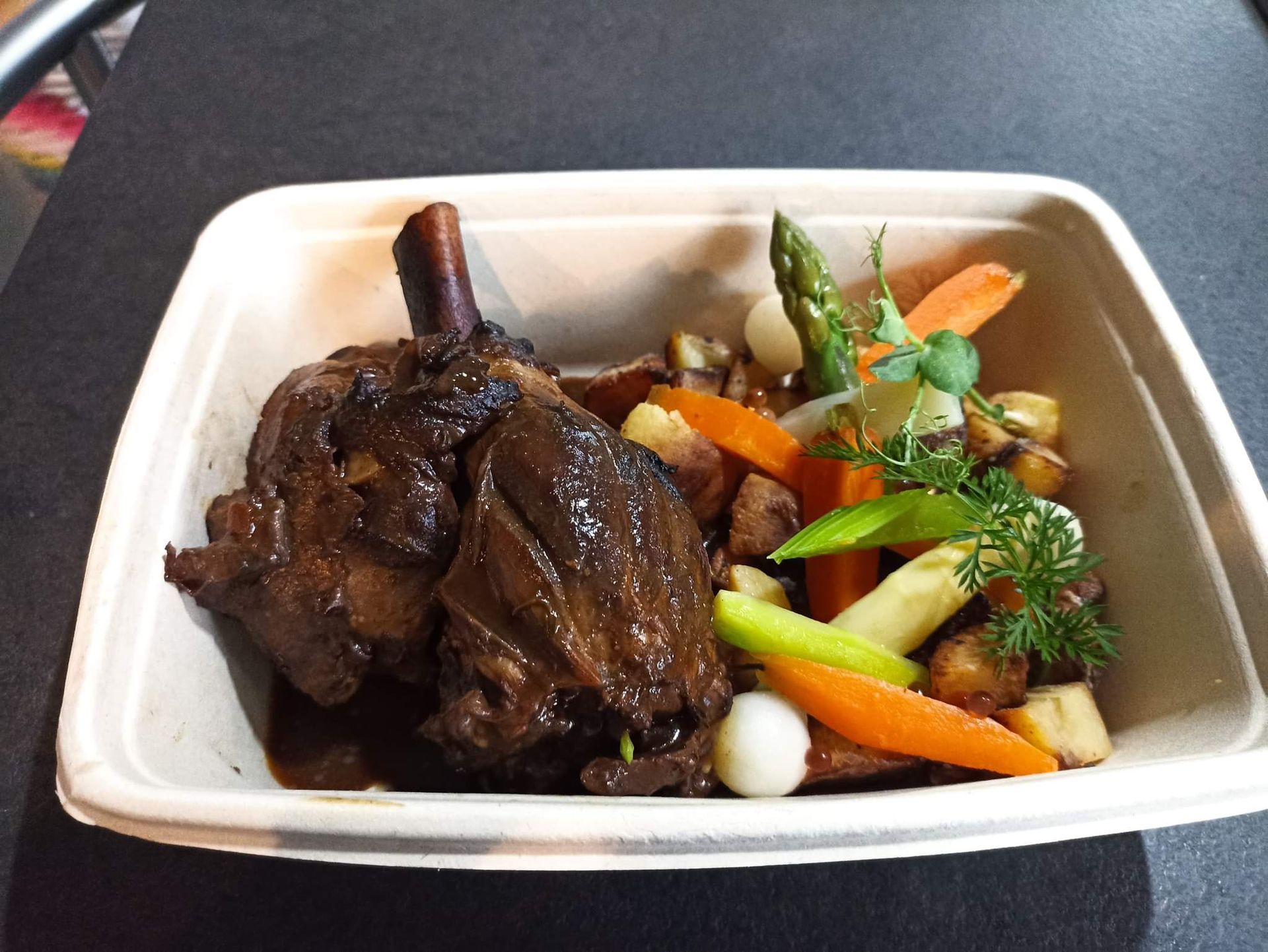 Barquette à emporter contenant de la viande braisée et des légumes variés, garnie d’herbes aromatiques