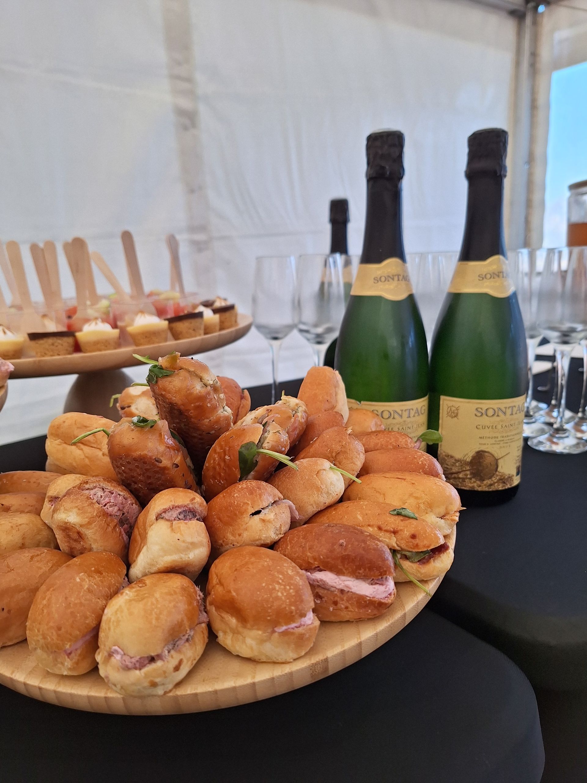 Des plateaux de pâtisseries dorées sur une table, avec deux bouteilles de vin pétillant, dans un espace événementiel sous chapiteau