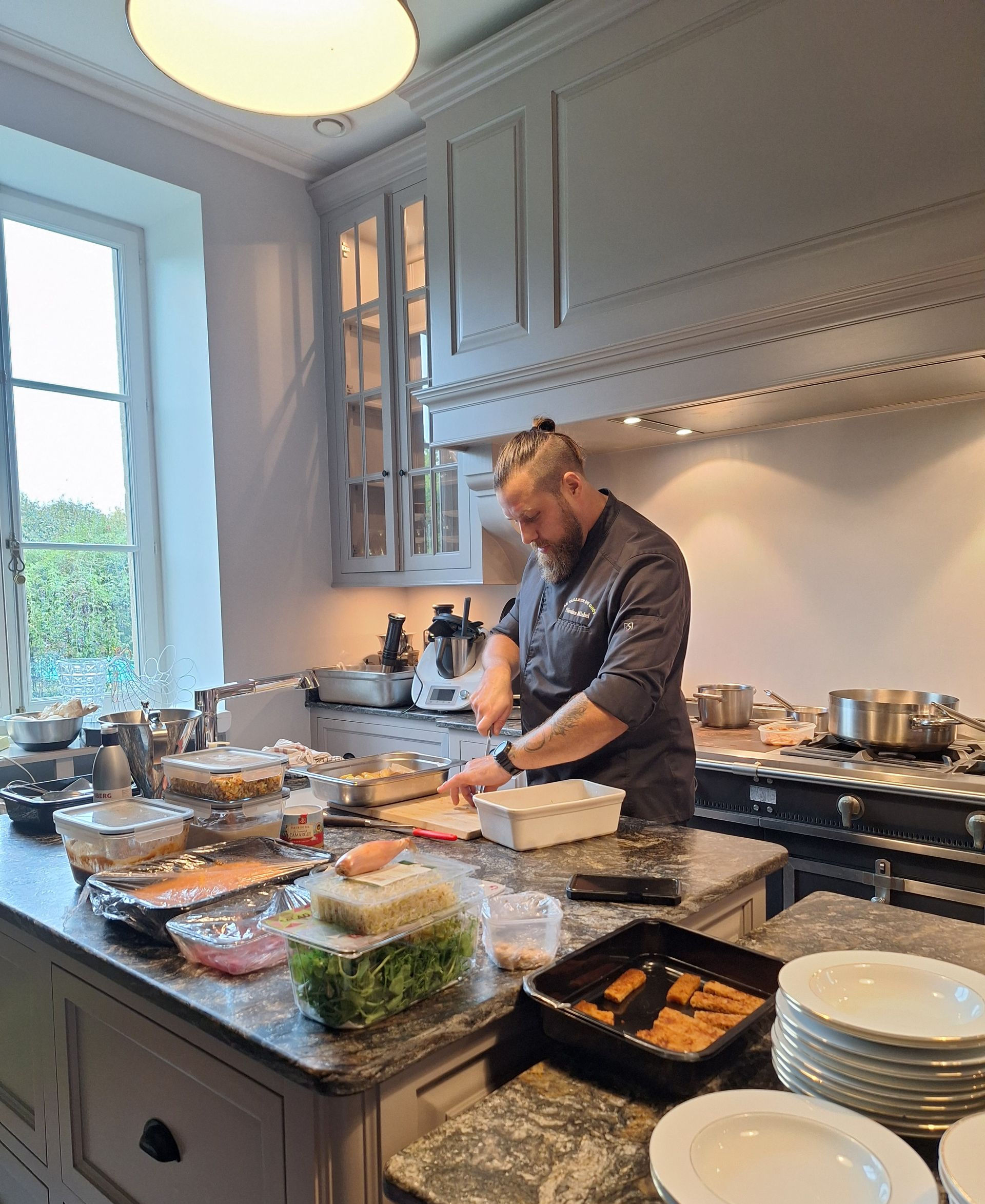 Un homme prépare à manger sur un îlot de cuisine, entouré de plats remplis d’ingrédients et d’assiettes empilées, dans une cuisine lumineuse