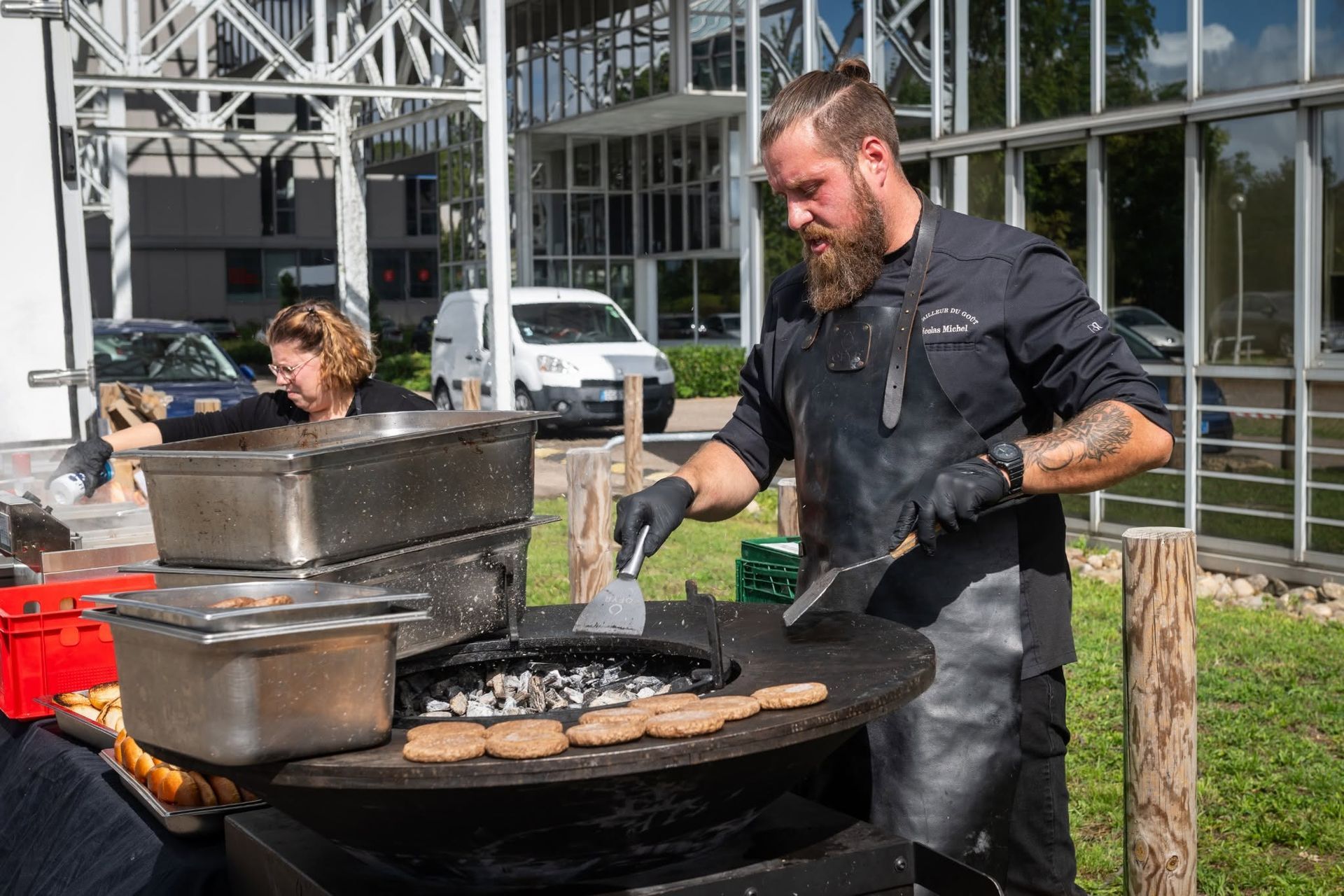 Un chef fait griller des burgers en plein air sur un stand de marché, avec une autre personne à proximité et un bâtiment en arrière-plan