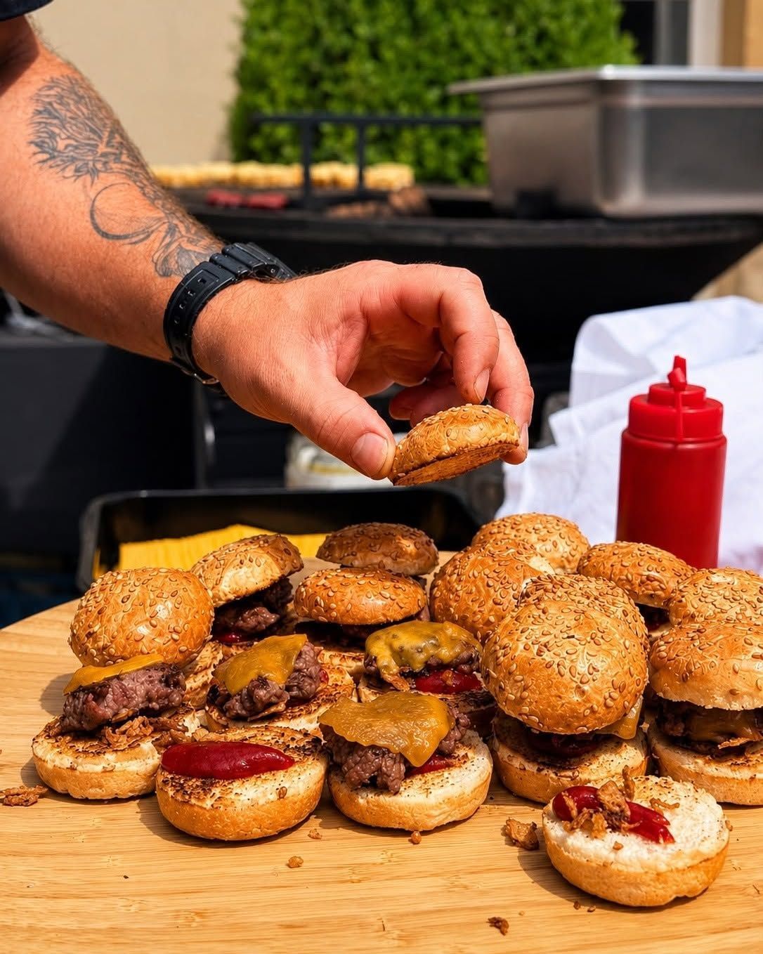 Une main dépose un pain à burger au sésame sur une pile de miniburgers, à côté de bouteilles de ketchup rouge, en plein air