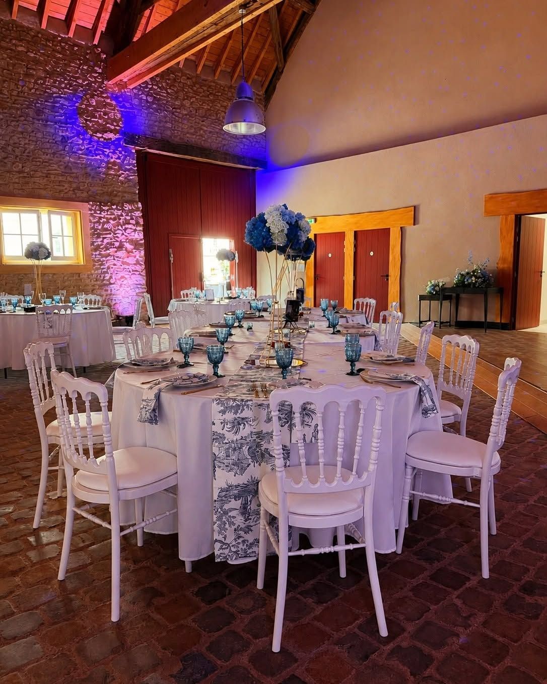 Table ronde de banquet dressée avec des chaises blanches et une décoration florale bleue, dans une salle de réception rustique à l’éclairage chaleureux