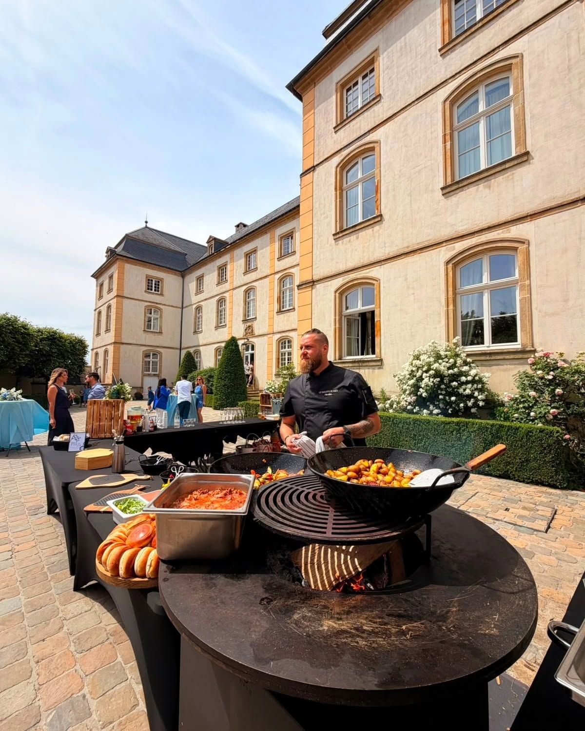 Buffet en plein air avec des plats disposés sur des grils ronds dans une cour, à côté d’un bâtiment beige, et des personnes rassemblées à proximité