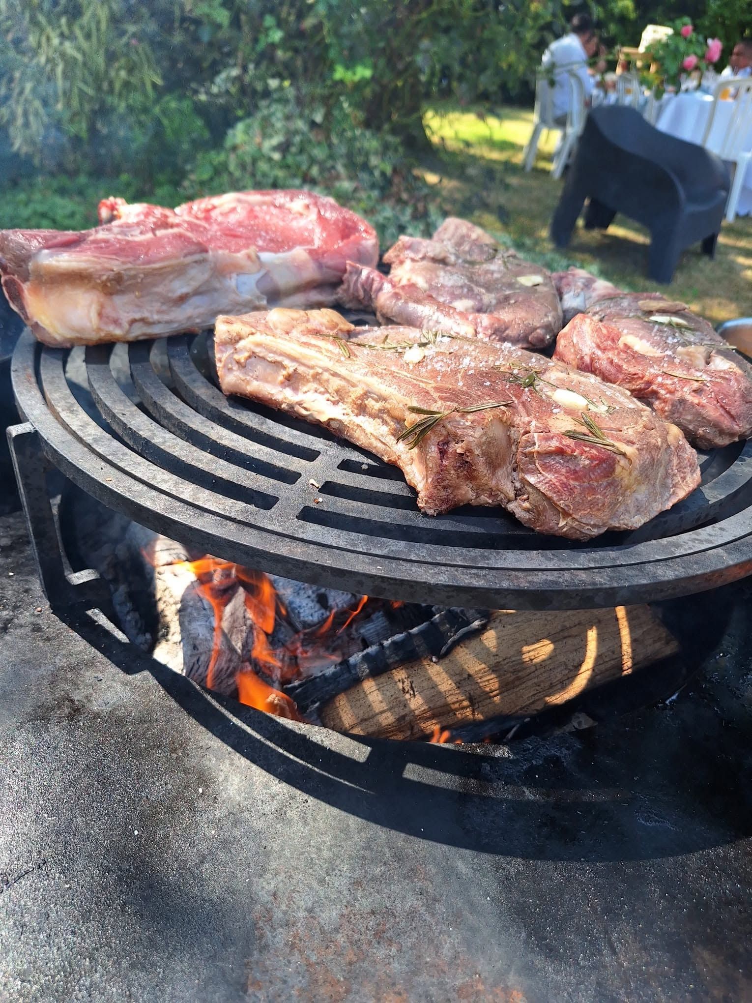 Des steaks et des côtes de bœuf grillent sur un barbecue rond au charbon de bois en plein air, avec des flammes visibles en dessous.