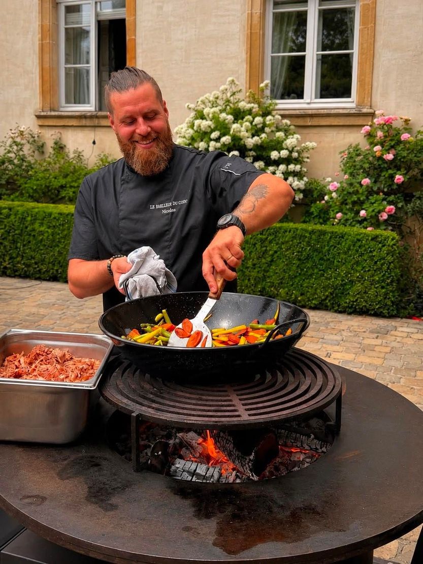 Un homme remue des aliments dans un wok au-dessus d’un feu de plein air, à côté de plateaux d’ingrédients, dans un jardin intérieur