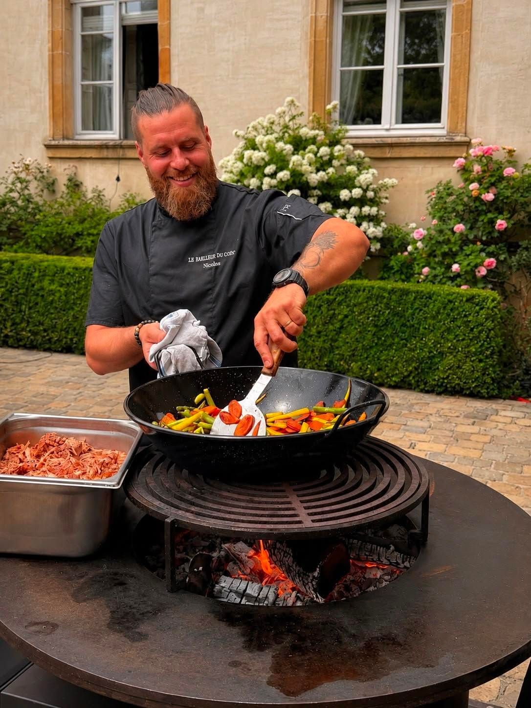 Un homme remue des aliments dans un wok au-dessus d’un feu de plein air, à côté de plateaux d’ingrédients, dans un jardin intérieur
