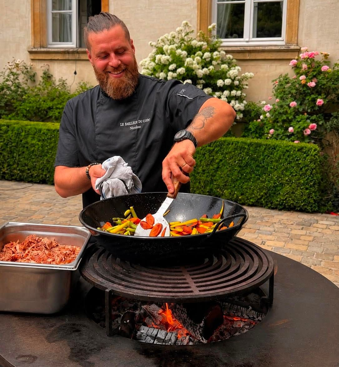 Un chef fait sauter des légumes dans un wok au-dessus d’un feu en plein air, avec des plateaux d’ingrédients à proximité