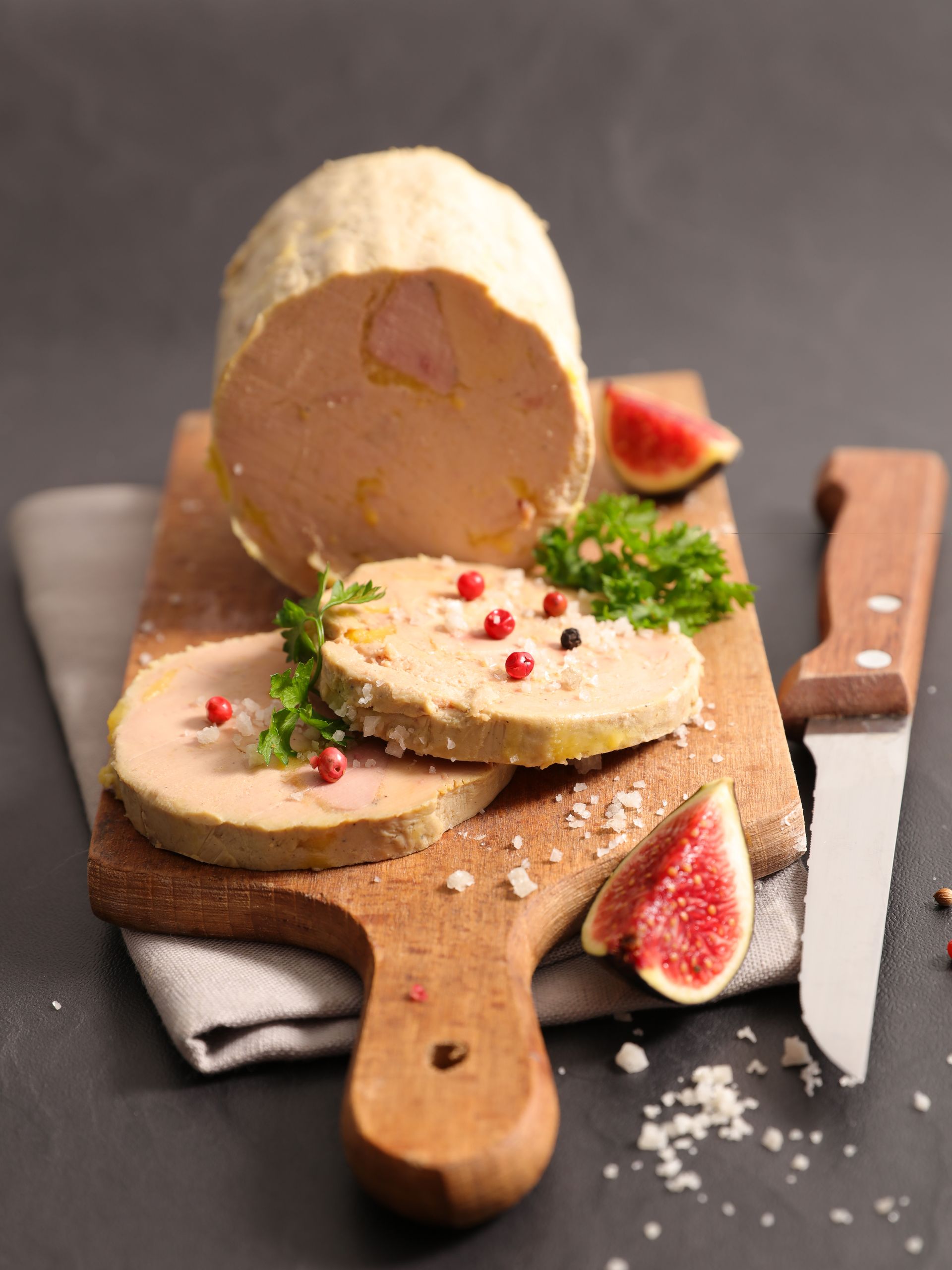 Bûche de foie gras sur une planche en bois, accompagnée de crackers, d’herbes aromatiques, de tranches de figue et d’un couteau, sur une table sombre
