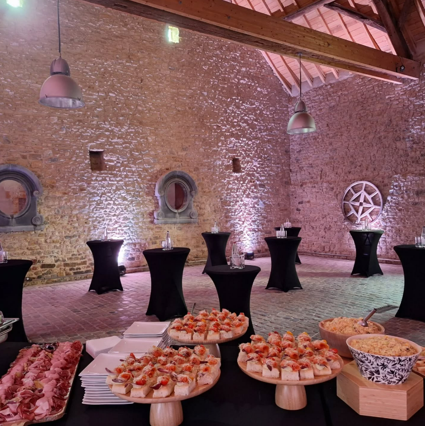 Buffet disposé sur des tables rondes dans une salle de réception rustique aux murs de pierre, avec des suspensions et des poutres apparentes