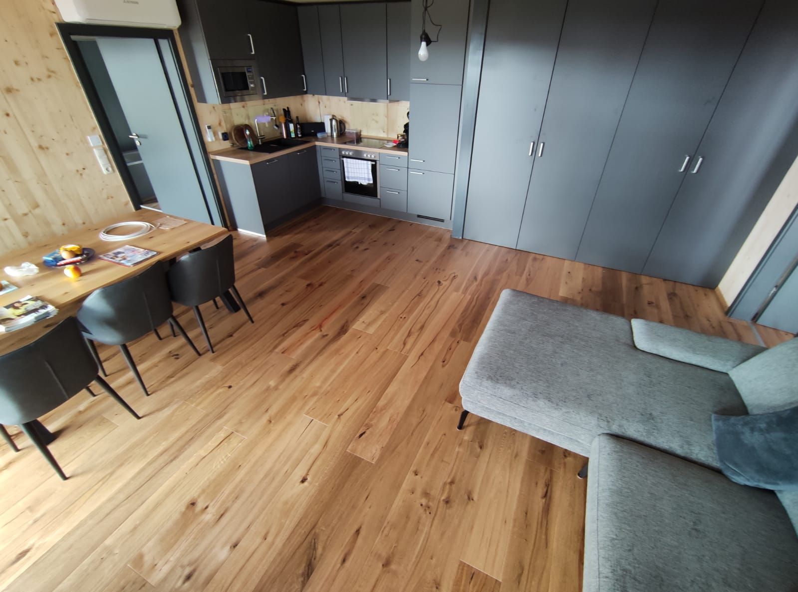 Wohnraum mit Holzboden, Küche, Esstisch mit Stühlen und einem grauen Sofa.