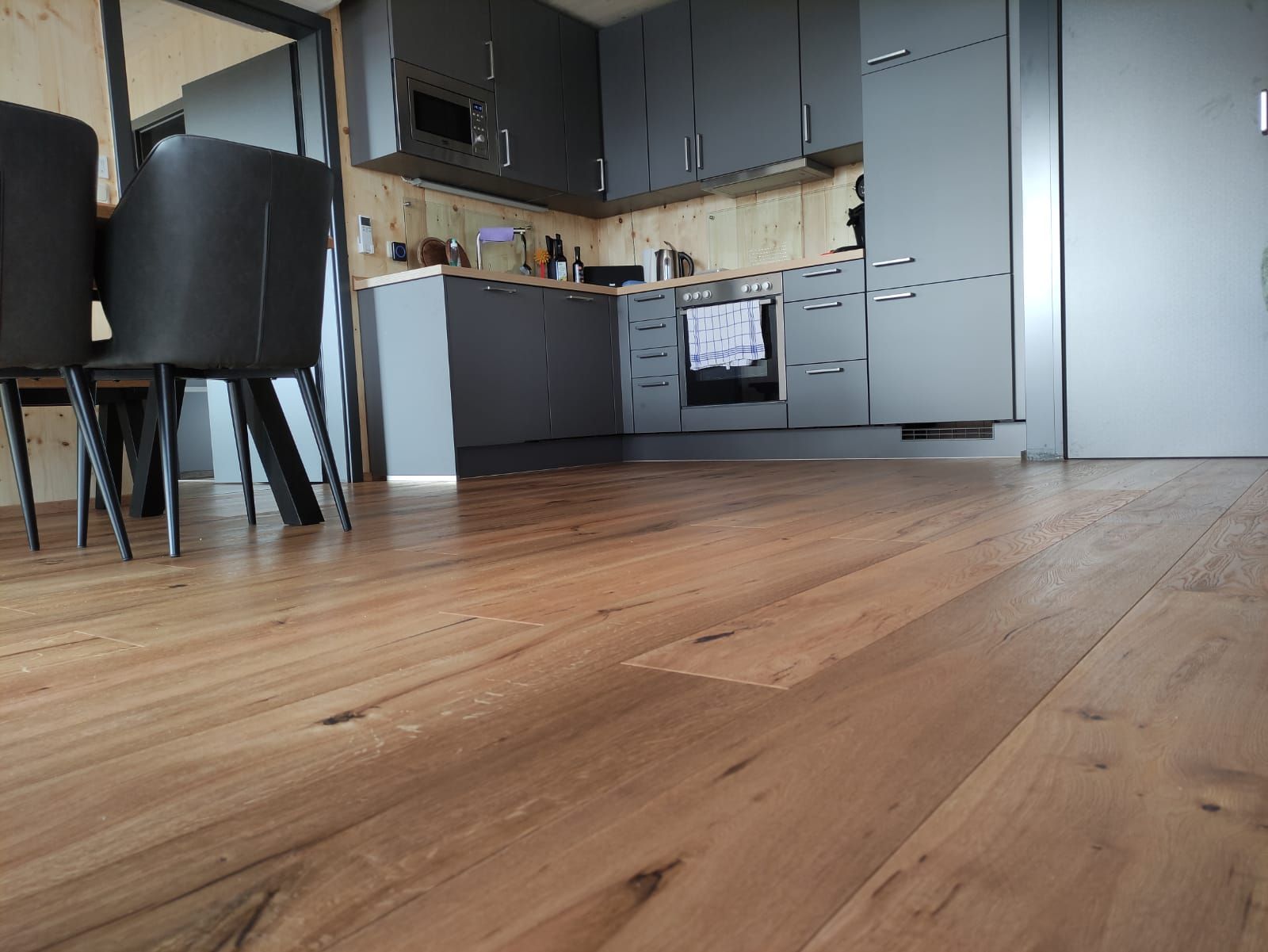 Holzboden, im Hintergrund eine Küche; graue Schränke und Geräte.