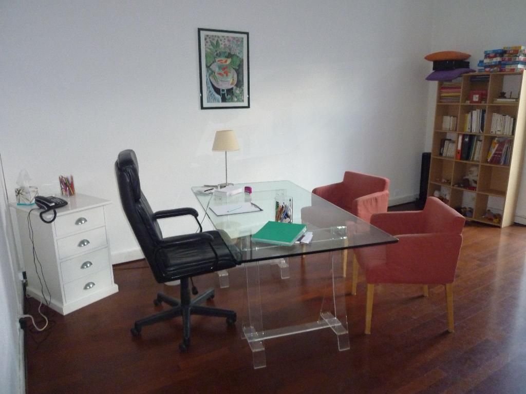 Bureau à domicile avec bureau en verre, chaise noire, fauteuils rouges, lampe et bibliothèque sur un parquet en bois.