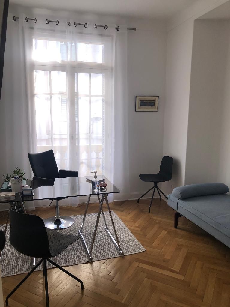 Salon lumineux avec une grande fenêtre, deux fauteuils, un tapis, une petite table et un canapé gris.