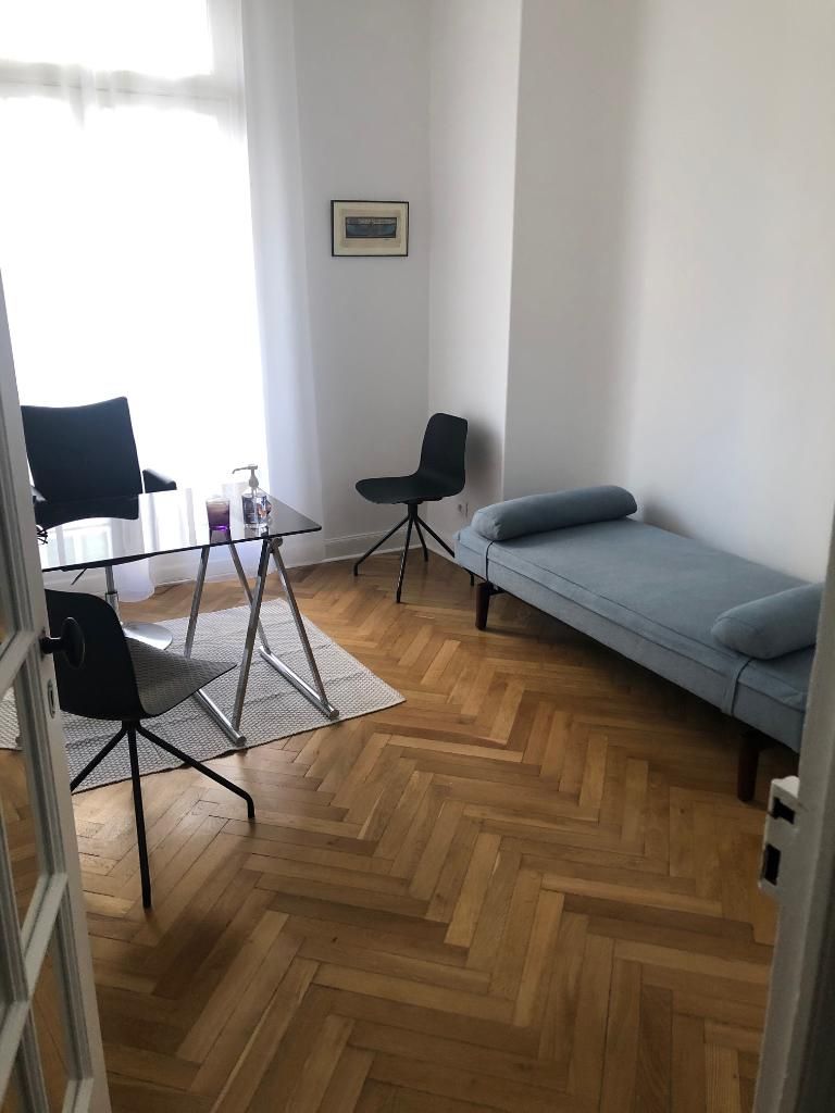 Salon minimaliste avec parquet en chevrons, canapé gris, chaises noires et petite table en verre près d’une fenêtre