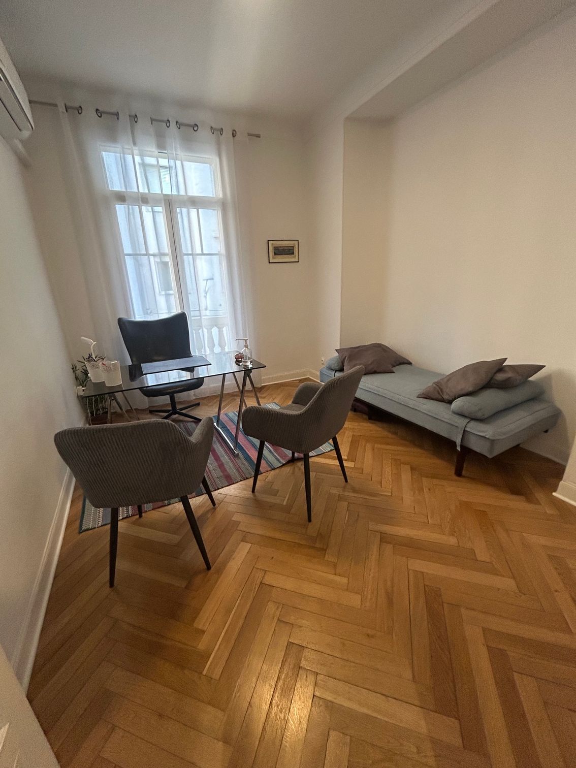 Salon lumineux avec une table et des chaises, un canapé gris et une grande fenêtre ornée de rideaux transparents.
