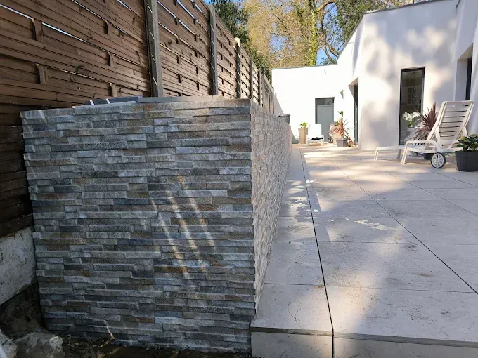 Mur de patio en pierre jouxtant une terrasse pavée dans une cour arrière, avec une clôture en bois et des chaises blanches en arrière-plan.