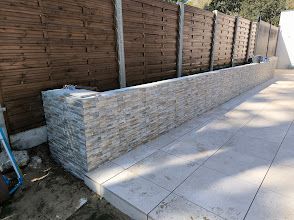 Muret en pierre bordant une terrasse pavée et une clôture en bois dans un jardin.