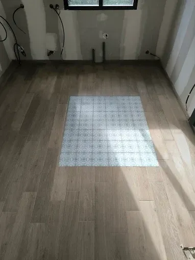 Pièce vide avec parquet clair et un tapis à carreaux blancs au centre, près d'une fenêtre.