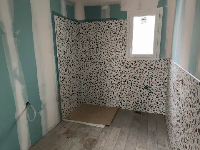 Petite salle de bains carrelée en cours de rénovation, avec murs à rayures, douche à motifs et fenêtre étroite.