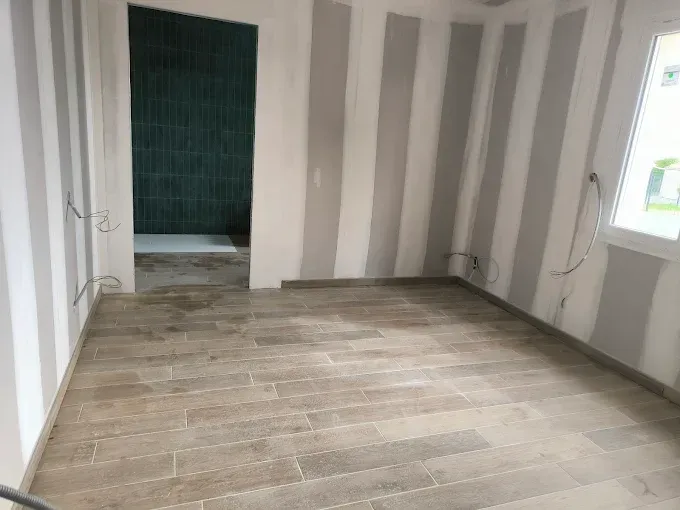 Pièce vide aux murs gris rayés, au sol imitation bois et avec une fenêtre sur la droite.