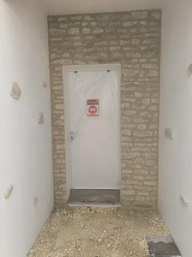 Couloir étroit et inachevé avec une porte en pierre et une porte blanche au fond, sol recouvert de gravats.