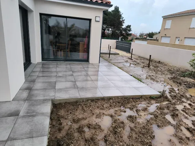 Terrasse extérieure carrelée de gris, jouxtant une maison, un jardin sablonneux et un muret.