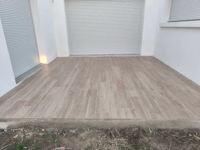 Patio carrelé vide avec sol imitation bois, volets blancs fermés et un petit éclairage d'angle.