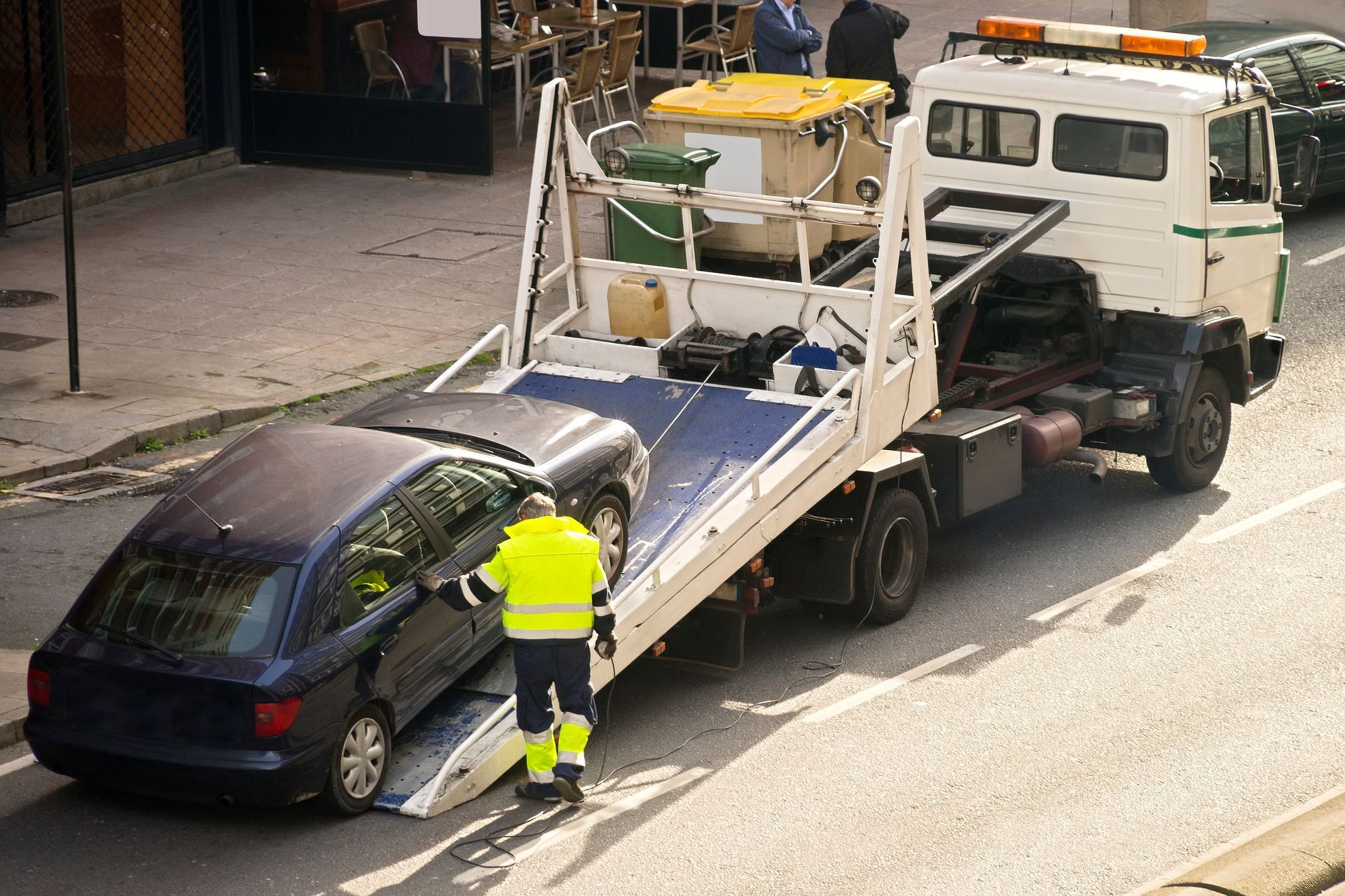 Une dépanneuse charge une voiture de couleur sombre dans une rue de la ville. Un ouvrier en gilet jaune guide le véhicule.
