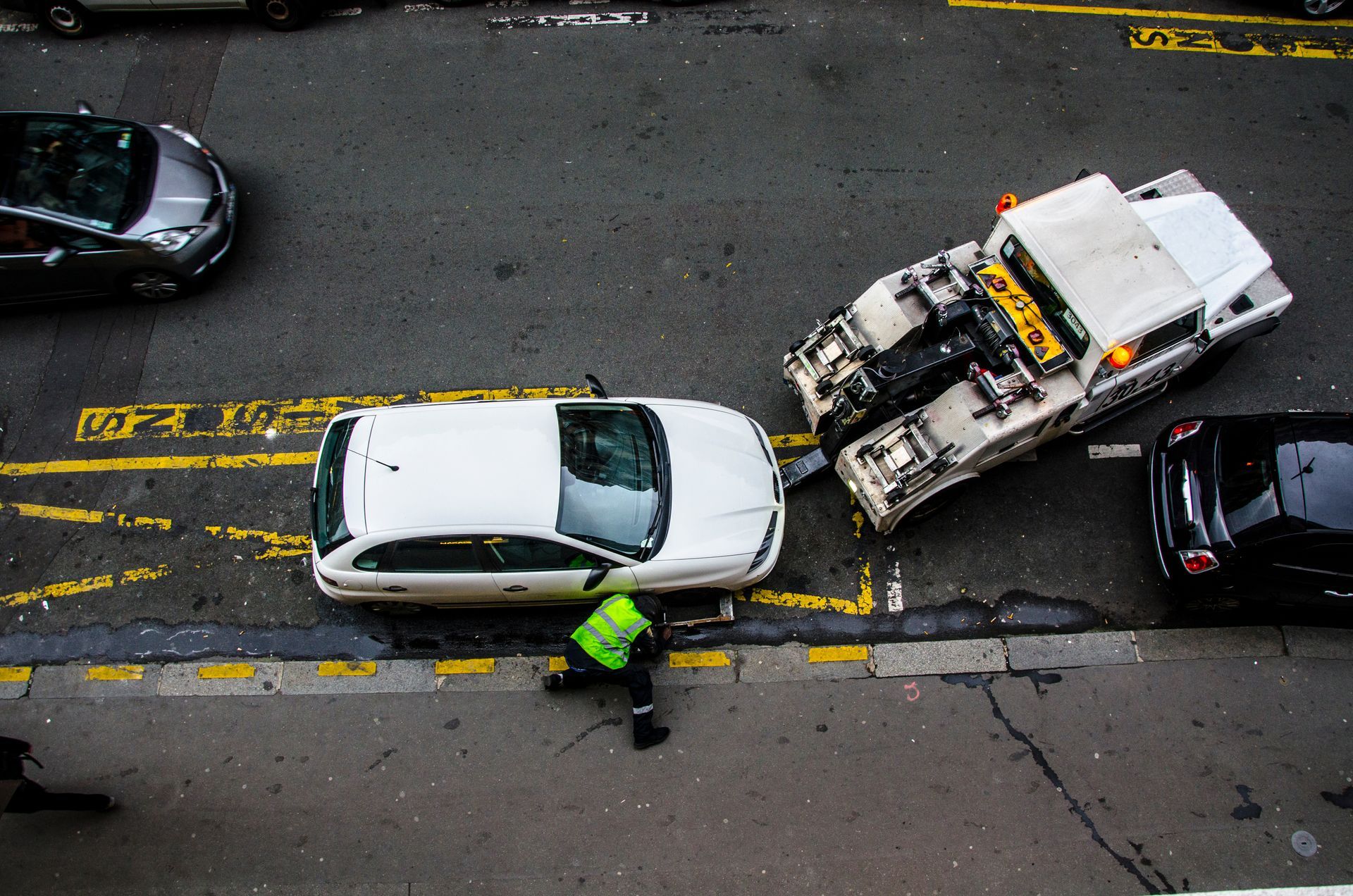 Une dépanneuse remorque une voiture blanche garée dans la rue, avec un agent de stationnement.