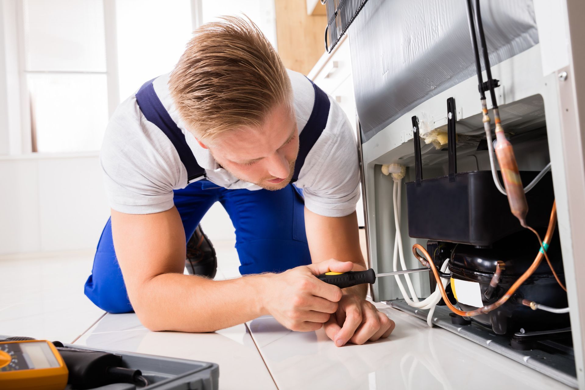 Un hombre está arrodillado en el suelo arreglando un refrigerador.