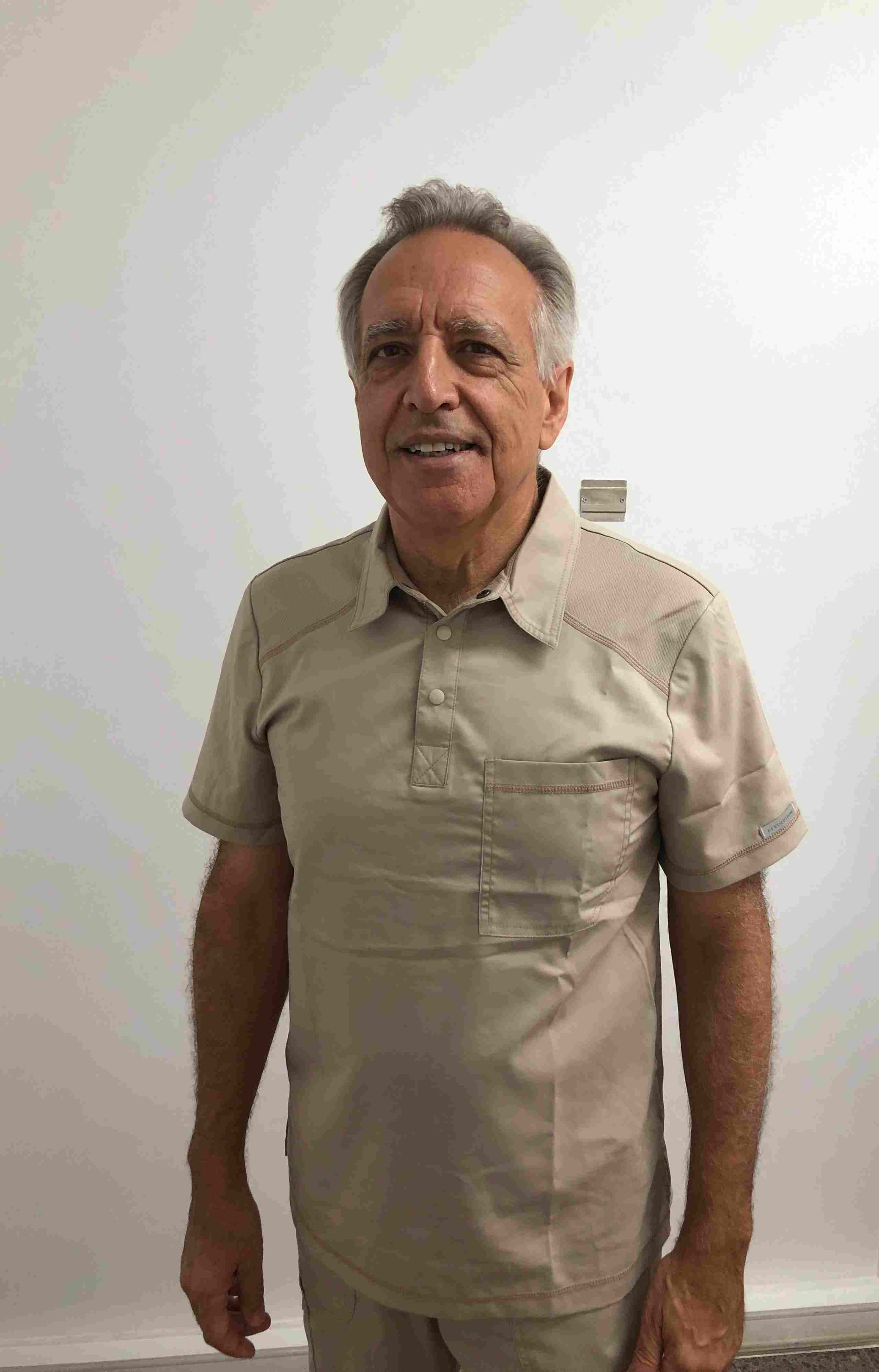 Un hombre con una camisa color canela y pantalones color caqui está parado frente a una pared blanca.