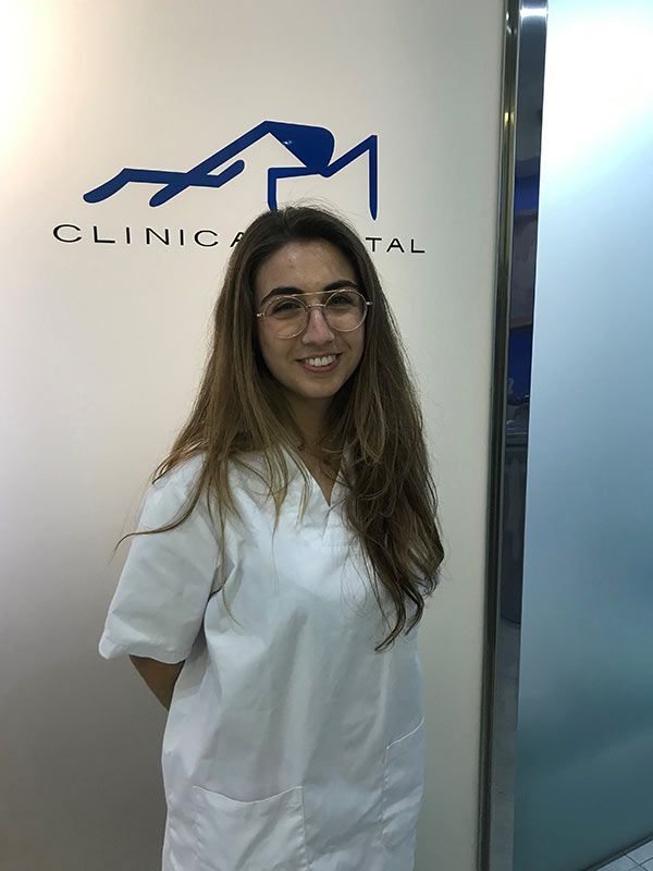 Una mujer que lleva gafas y una bata blanca está parada frente a un cartel que dice Clínica Dental.