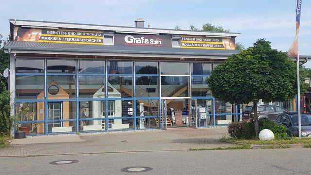 Ein modernes Geschäftsgebäude mit großen Glasfenstern und einem Haupteingang, auf dessen Dach das Schild „Graf & Sohn“ prangt.