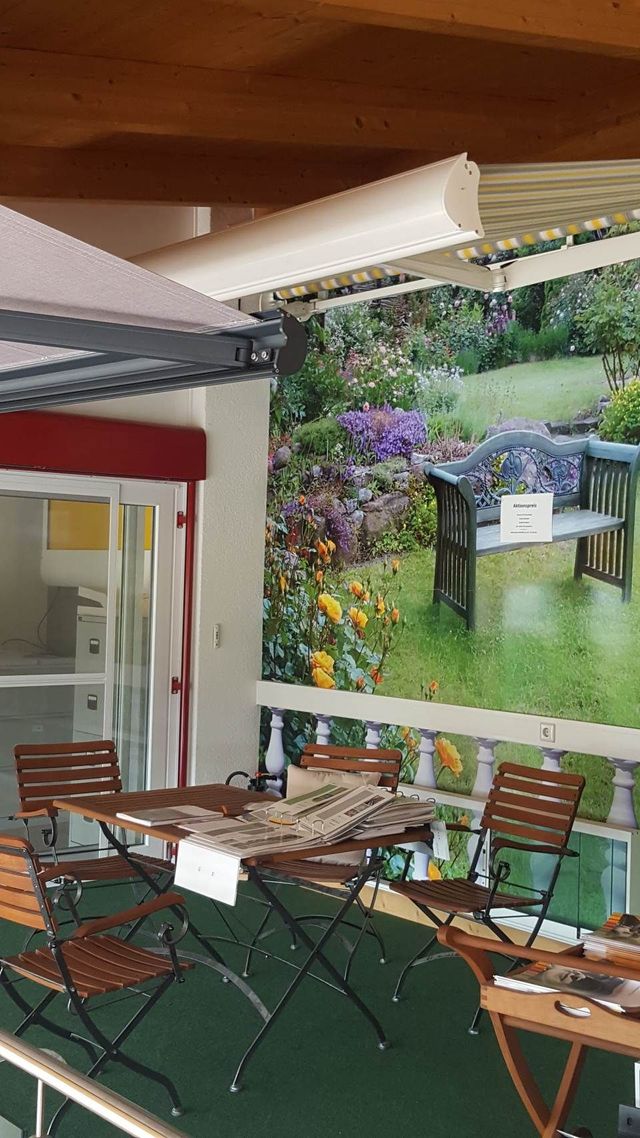 Eine Terrasse mit braunen Metallmöbeln auf grünem Boden, an deren Wand ein großes Wandtattoo eine Gartenszene mit einer Holzbank zeigt.