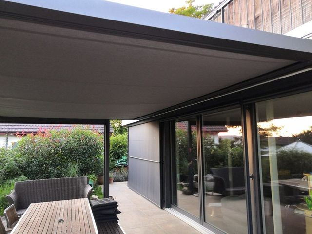Eine moderne Terrasse mit einziehbarem Vordach, Gartenmöbeln und großen Glasschiebetüren zum Haus.