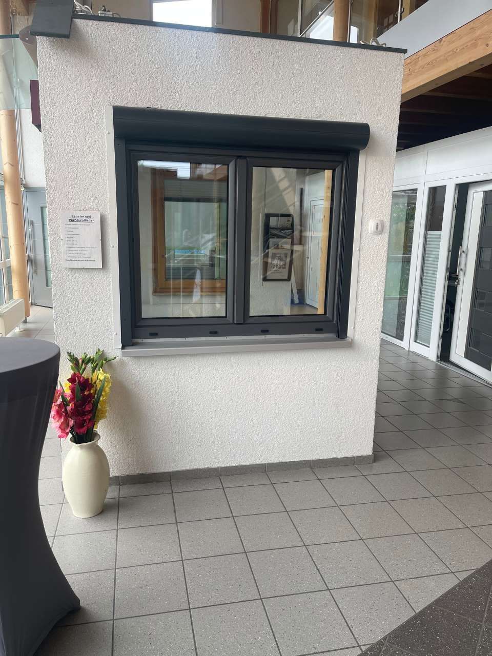 Eine weiße, strukturierte Gebäudewand mit einem dunkel gerahmten Fenster, einem eingebauten Fensterladen und einer Blumenvase auf dem gefliesten Boden.
