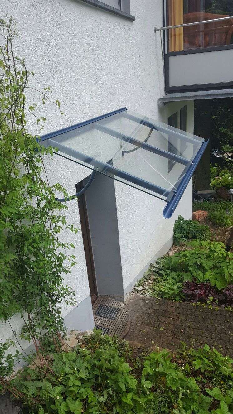 Ein modernes Vordach aus Glas und dunklem Metall, das über einer Seitentür an einer weißen Stuckaußenwand angebracht ist.