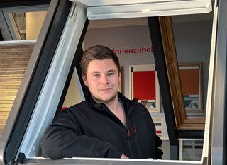 Eine Person in einer schwarzen Jacke lehnt sich aus einem Fensterrahmen; im Hintergrund sind Muster von Fensterjalousien zu sehen.