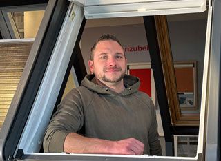 Eine lächelnde Person in einem olivgrünen Kapuzenpullover lehnt sich durch ein offenes Oberlicht in einem Ausstellungsraum.