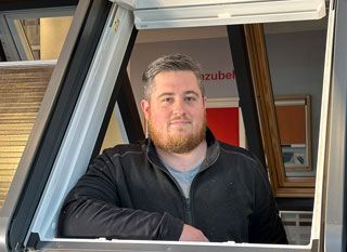 Eine Person mit Bart und dunkler Fleecejacke lehnt sich aus einem offenen Dachfenster in einem Ausstellungsraum.