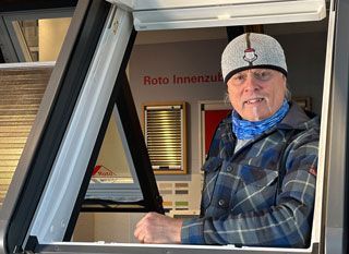 Eine Person mit Mütze und blau gemustertem Schal lehnt sich durch ein offenes Dachfenster in einem Zimmer mit Jalousien.