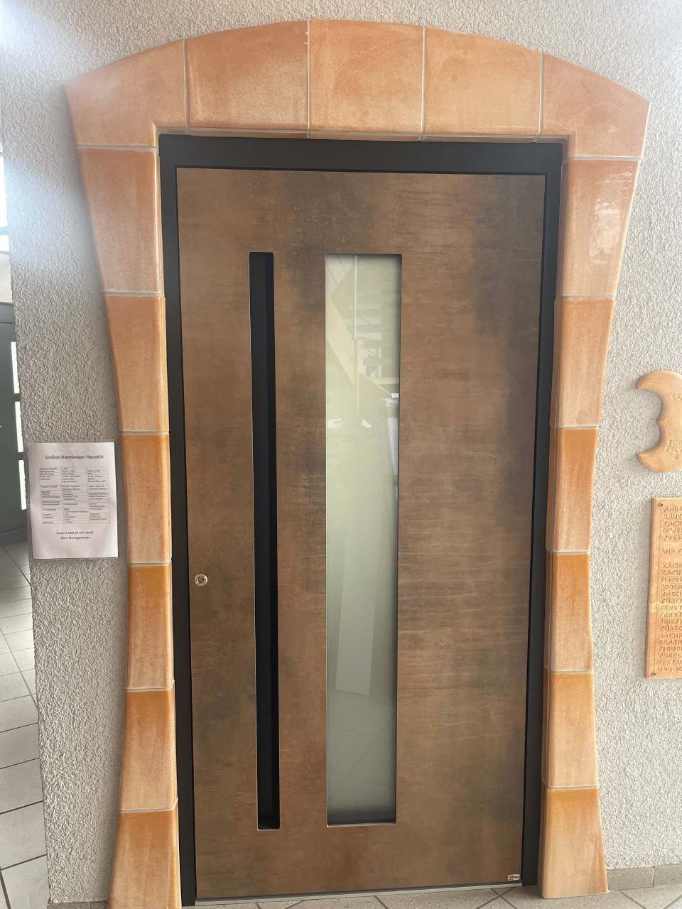 Eine Holztür mit einer vertikalen Glasscheibe, die in einen gewölbten, mit Steinfliesen verkleideten Rahmen an einer strukturierten Wand eingefasst ist.