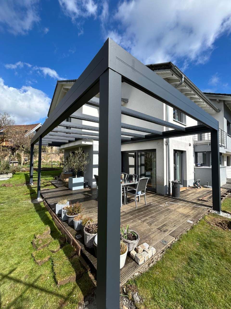 Unter einem teilweise bewölkten blauen Himmel steht eine moderne, anthrazitgraue Pergola über einer Steinterrasse neben einem Haus.