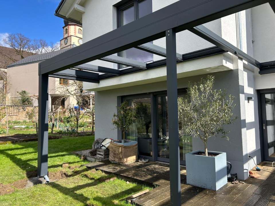 Moderne Pergola aus schwarzem Metall über einer Holzterrasse mit Olivenbäumen in Töpfen neben einem weißen Haus.