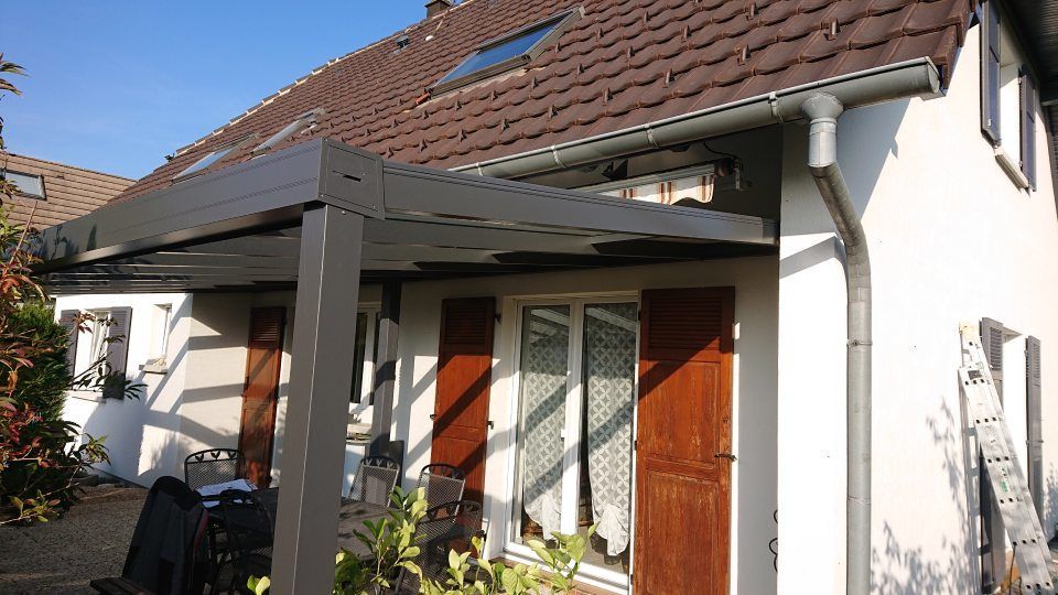 An einem sonnigen Tag lehnt eine graue Pergola an der Seite eines zweistöckigen Hauses mit braunen Fensterläden und Ziegeldach.